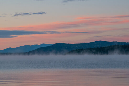 Pink sunrise over mountains and calm lake with mist rising from water