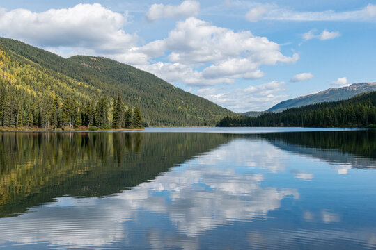 Calm lake between mountains and forest with reflections of puffy clouds in blue water