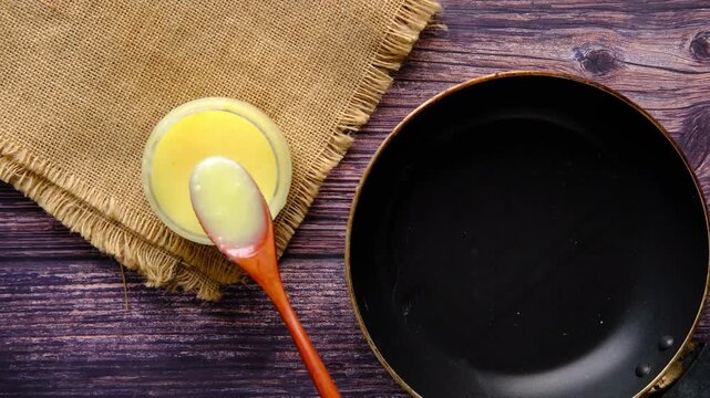 A hand using a wooden spoon to scoop ghee from a small glass bowl and transfer it into a black pan on a wooden table with burlap cloth, top view, cinematic footage