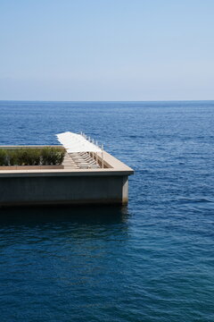 Modern seaside deck with a row of white sun loungers and umbrellas overlooking a vast blue sea under a pale, cloudless sky.