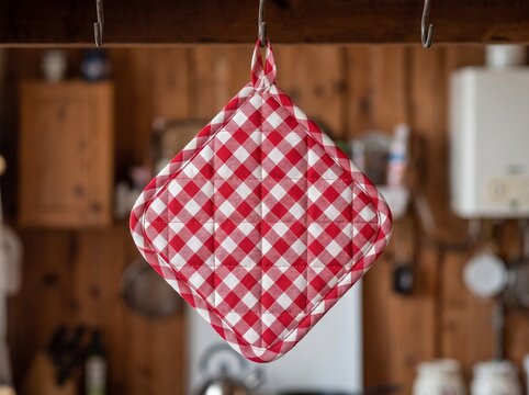 Red and white gingham checkered quilted pot holder hanging from a metal hook in a rustic wooden kitchen with blurred background