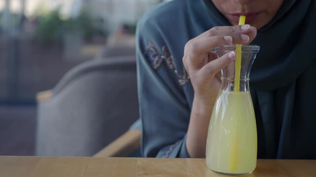 Woman in hijab drinking fresh lemonade through yellow straw in glass carafe at outdoor cafe terrace