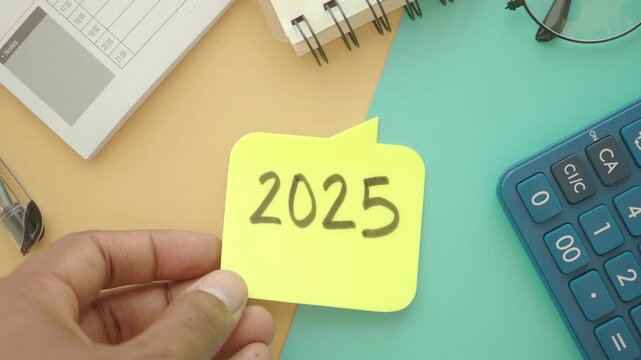 Hand holding yellow sticky note with 2025 written on it next to calculator and notebook