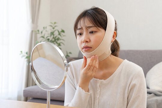 顔の脂肪吸引後に圧迫バンドを着け鏡で確認する女性