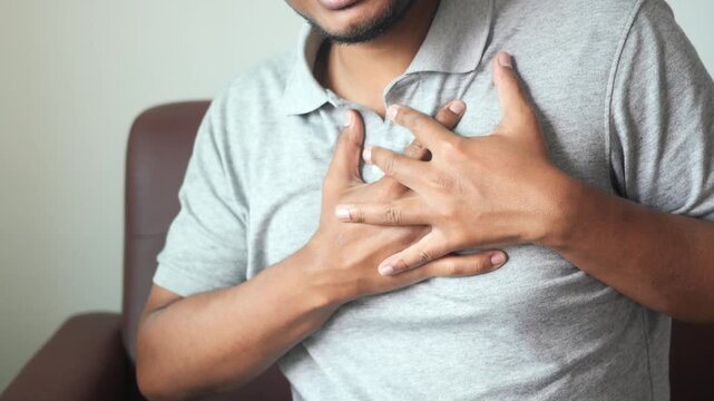 Unrecognizable young man experiencing sharp chest pain and heart attack while sitting on sofa at home indoors