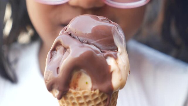Young girl in pink glasses taking bite of melting chocolate and vanilla ice cream cone outdoors