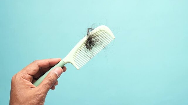 A male hand holds a white plastic comb with a clump of black hair and then removes the hair against a light blue background, showcasing motion and the issue of hair loss.