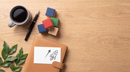Fototapeta premium Business Startup Launch Concept with Rocket Drawing on Notebook Beside Coffee Cup Pen and Wooden Blocks on Wood Table Background for Success Strategy Planning and Creative Growth Vision Top View