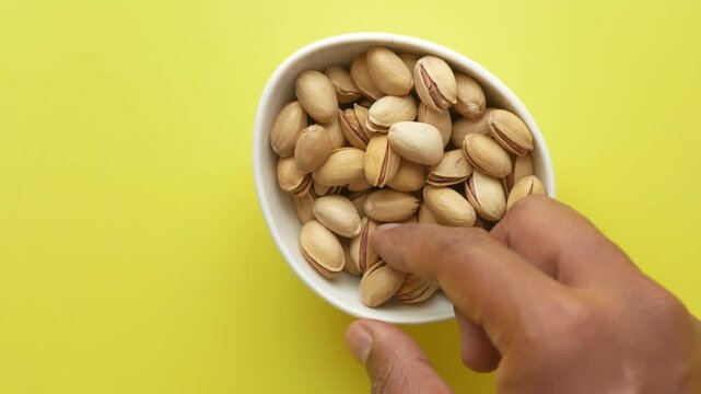 A male hand takes one pistachio nut from a bowl of pistachios on a yellow background, cinematic close-up shot of a healthy snack