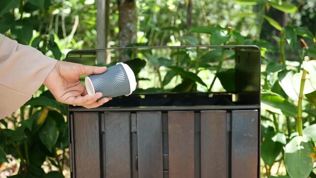 Person hand throwing paper cup into outdoor trash bin, green foliage background