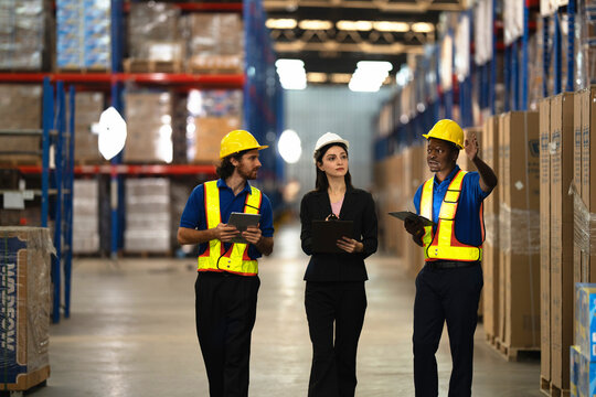 Logistics team standing between tall pallet racking, the warehouse worker gesturing up at a high storage bay while his manager and teammate keep the order list open