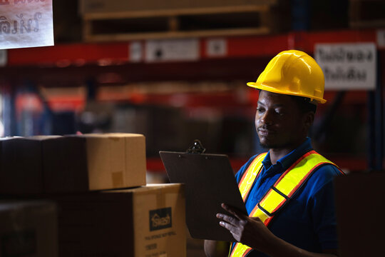 Warehouse operative smiles faintly as he reads his clipboard beside stacked cardboard shipping boxes in the low lit storage aisle of a logistics distribution centre