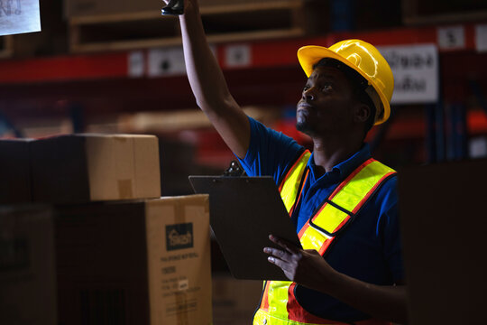 Warehouse employee scanning barcodes on the upper shelf of blue and orange pallet racking while holding a clipboard in a storage facility, working under low warehouse light