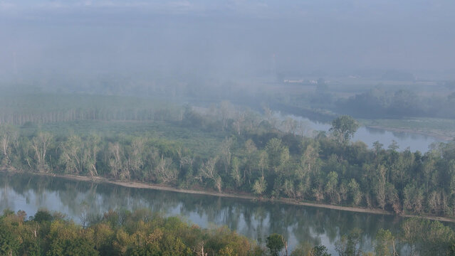 Po River winding through Monticelli d'Ongina landscape, dense fog and mist lingering over water and trees, reflecting an overcast sky after a hailstorm in Emilia Romagna
