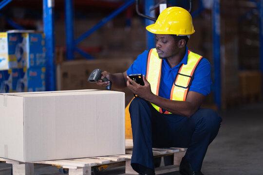 African warehouse worker in a blue polo and reflective vest crouches beside a large cardboard box on a wooden pallet holding a barcode scanner and handheld terminal