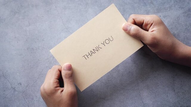 Top view of hands holding beige thank you card on grey background with subtle movement for message of gratitude