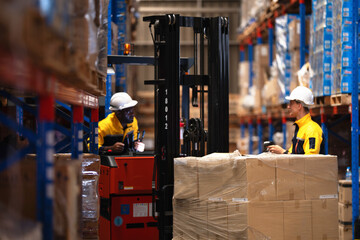 Wide warehouse aisle shot as a forklift operator holds a scanner in the cab and his teammate reaches a hand to the stretch wrapped pallet of cardboard boxes beside him © ultramansk