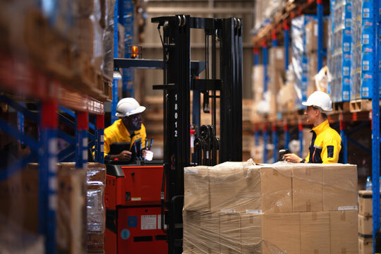 Wide warehouse aisle shot as a forklift operator holds a scanner in the cab and his teammate reaches a hand to the stretch wrapped pallet of cardboard boxes beside him
