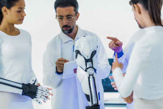 Artificial intelligence research team at a benchtop robot arm: a colleague points out a control detail while the lead engineer adjusts the manipulator in a white lab
