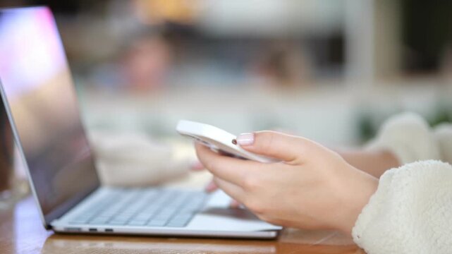 Hands hold phone near laptop. Soft focus background. Warm indoor lighting. Person wears cozy sweater. Scene shows multitasking with devices. Focus on hands and technology use