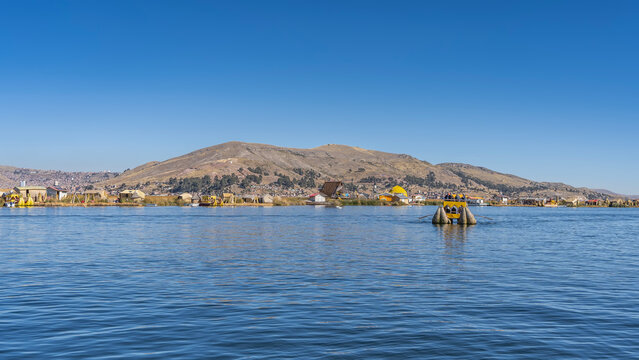 A traditional balsa de totora boat carrying tourists is sailing across a high-altitude lake. On the shore, there are reed huts and pavilions. Mountains against a blue sky. Peru. Puno. Lake Titicaca.

