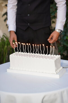 White celebration cake with lit candles on a table, while a man in a classic black suit stands behind it.