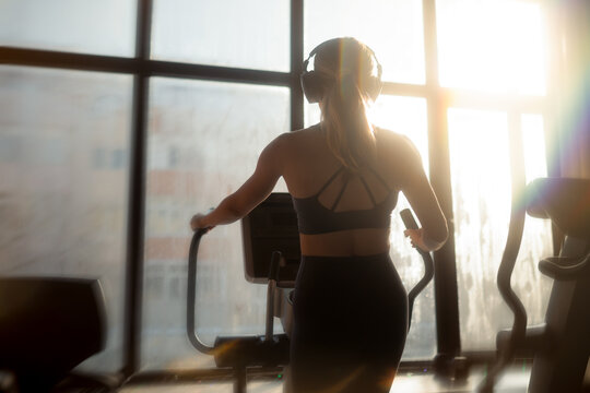 Back View Of Athletic Woman Exercising On Elliptical Gym Machine. Female athlete with headphones training on cardio cross trainer in front window daylight