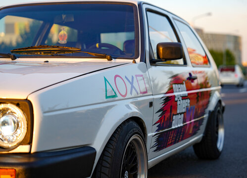 Berlin, Germany - May 24, 2024: Custom white vintage hatchback car parked on an urban street during golden hour, decorated with gaming graphics and vinyl livery wrap. GTA concept.