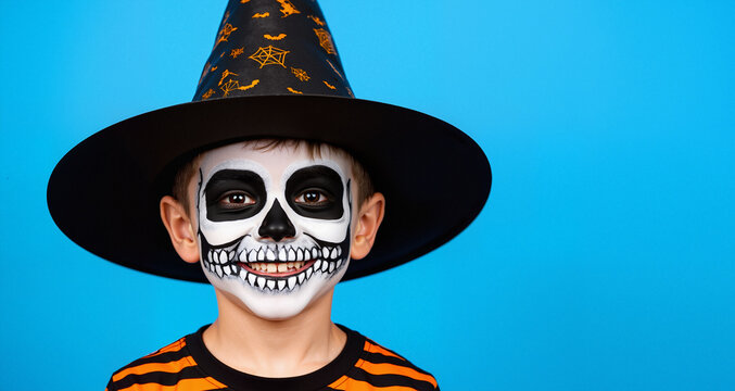 Smiling boy in skeleton face paint with witch hat, Halloween child with skull makeup on blue background, Fun Halloween portrait with copy space
