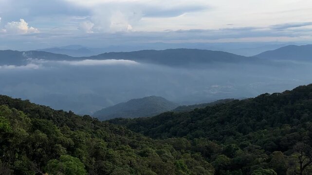 A panoramic view of lush green mountains under a cloudy sky. Mist covers the valleys, creating a serene and tranquil atmosphere.