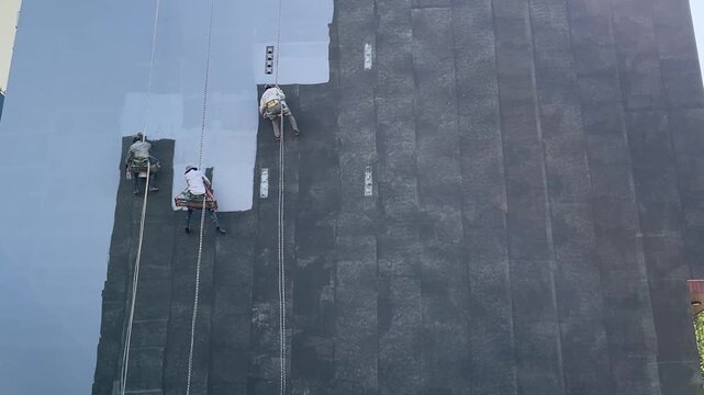 Three workers applying a coating to a large roof. They are using ropes for safety. The roof is partially blue and gray. Bright sunlight is present.