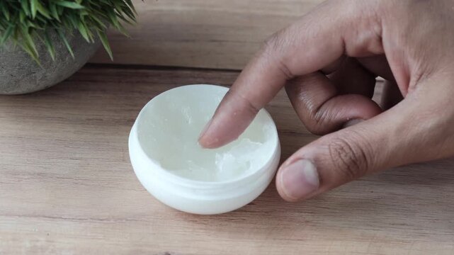 Hand taking white petroleum jelly from small plastic container on wooden surface near green plant