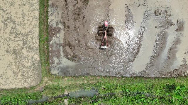 An aerial view of a Asian farmer operating a hand tractor to plow a muddy rice field prepare the soil for agricultural planting under bright natural sunlight in a rural traditional countryside.