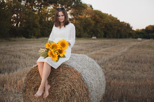 woman in a white dress holding a bouquet of wild flowers with sunflowers in a mowed field with straw bales