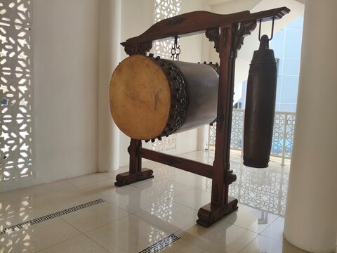 A traditional Indonesian Bedug drum and hanging wooden instrument called " kenthong" displayed on a wooden stand inside a brightly lit mosque with decorative latticework.