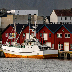 Traditional red wooden fishing huts and white boat in a scenic nordic harbor beneath a steep dark mountain wall with copy space © Quality by Simon