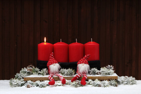 Four red Advent candles with two small gnome figures and fir branches on a snow-covered surface in front of a wooden wall with space for text. One candle is lit for the first Sunday of Advent.