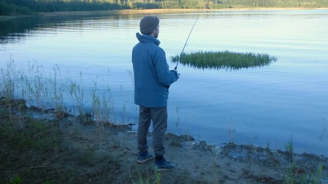Back view of young man wearing beanie and jacket standing on sandy shore of tranquil lake, casting fishing lure with spinning rod into calm morning water. Fisherman casting spinning rod into lake.