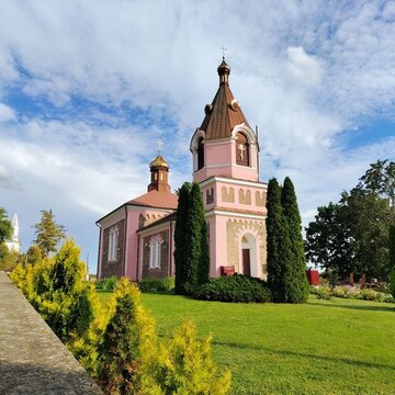 Beautiful pink Orthodox church with a bell tower, green lawn and tuya trees under a blue sky