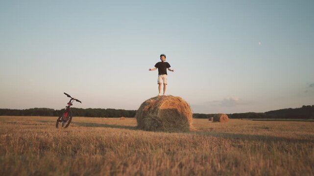 Carefree summer vacation in countryside, teen boy standing on haystack in field. Rest after long bike trip in nature, happiness and joy, positive emotions and active lifestyle in holidays, be happy