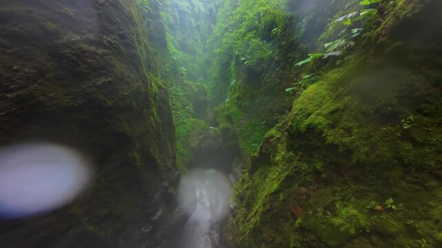 FPV drone flies low through lush mossy canyon in Costa Rica following turquoise river rapids waterfalls ferns and dense tropical rainforest walls with mist and sunlight breaking through leaves.