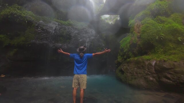 FPV footage in Costa Rica of a man standing with open arms before a misty jungle waterfall, surrounded by mossy cliffs, lush rainforest and a turquoise pool in a tropical canyon.