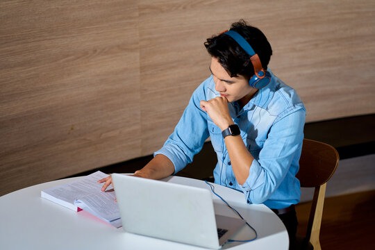 Focused young Asian man wearing headphones working on laptop and studying book at table in modern office. Student or freelancer attending online webinar.