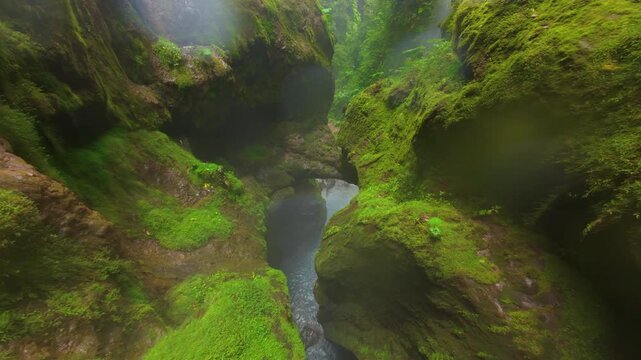 FPV drone flies through narrow mossy gorge toward turquoise river where father and son enjoy resting under waterfall in Costa Rica rainforest with mist rising over wet rocks and ferns.