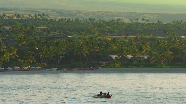Drone view of outrigger canoe with fishermen paddling calm ocean water at sunrise off Big Island in Hawaii with palm trees sandy beach and resort homes glowing in warm golden light.