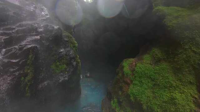 FPV drone flight into narrow misty canyon in Costa Rica with tall waterfall falling into turquoise pool surrounded by mossy volcanic rock walls and lush tropical rainforest plants.