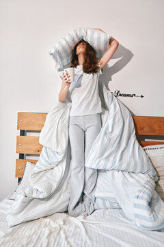 A tired woman holding a pillow and a mug in bed. She feels exhausted and overwhelmed. Concept for morning fatigue, insomnia, sleep issues, and stress.