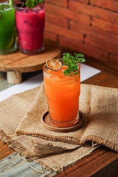 Fresh carrot iced beverage with mint and dried lime in a glass on wooden coaster over burlap, cafe background with brick wall.