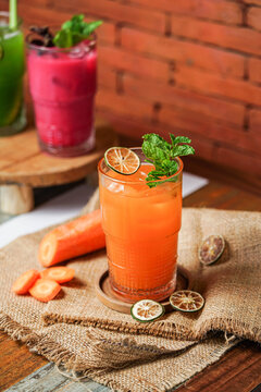 Fresh carrot iced beverage with mint and dried lime in a glass on wooden coaster, surrounded by carrot on burlap, cafe background with brick wall and other drinks.