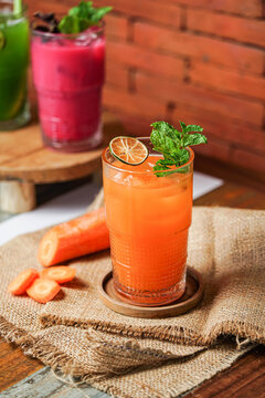 Fresh carrot iced beverage with mint and dried lime in a glass on wooden coaster, surrounded by carrot and citrus on burlap, cafe background with brick wall.
